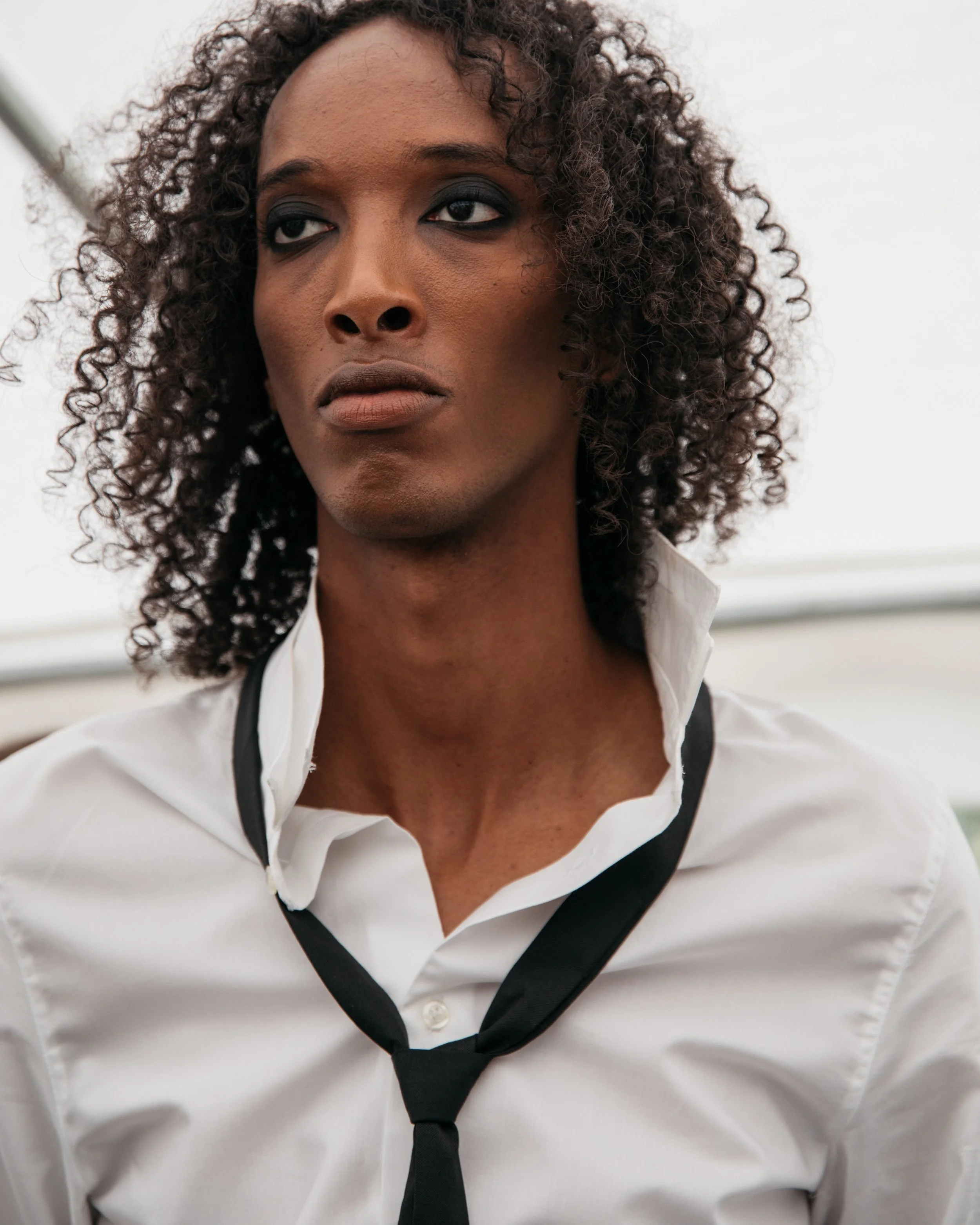 Close-up of a person with dark skin, curly hair, wearing a white shirt with the collar popped and a loosely tied black tie, looking contemplative.