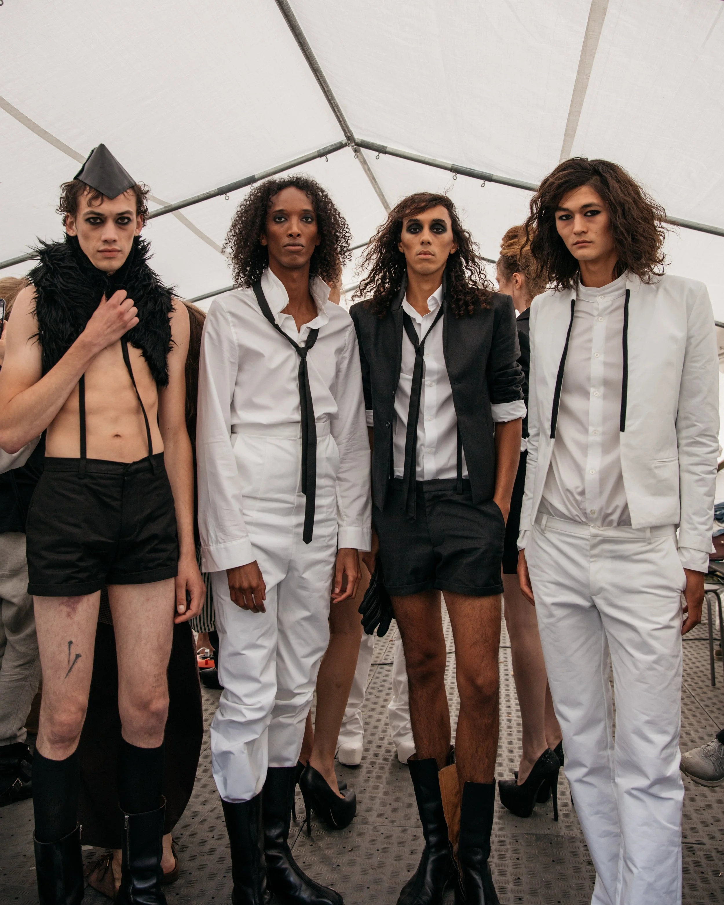 Group of four models standing in a tent, wearing black and white fashion outfits with dramatic makeup.