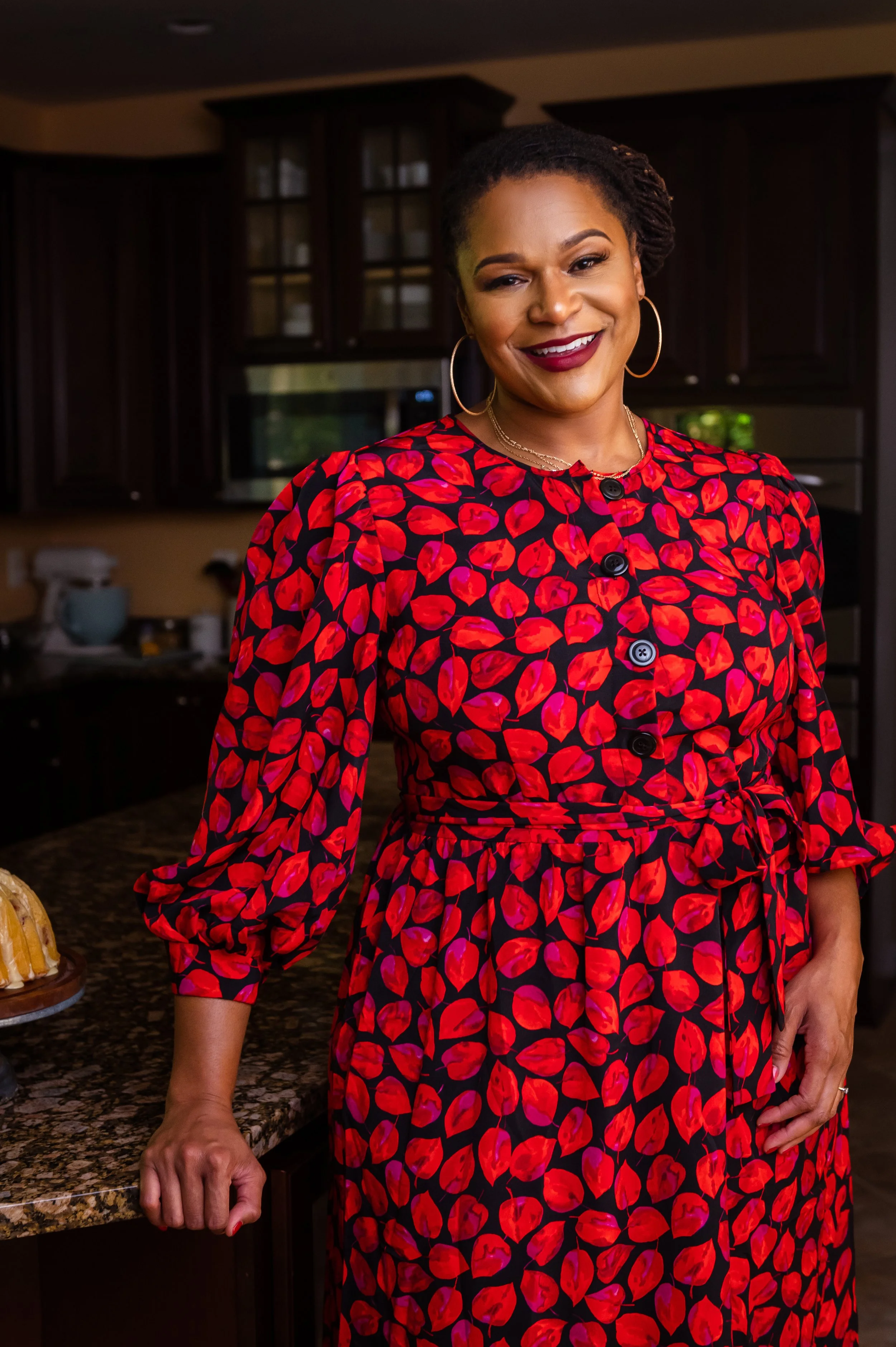 A woman smiling in a kitchen, wearing a red and black patterned dress with long sleeves and buttons, standing next to a granite countertop.
