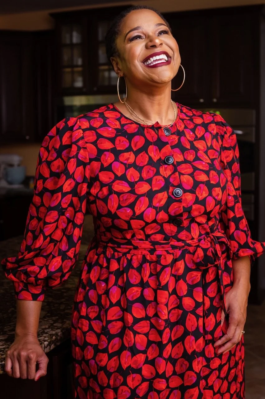 A joyful woman wearing a red and black patterned dress with large buttons, standing inside a kitchen.