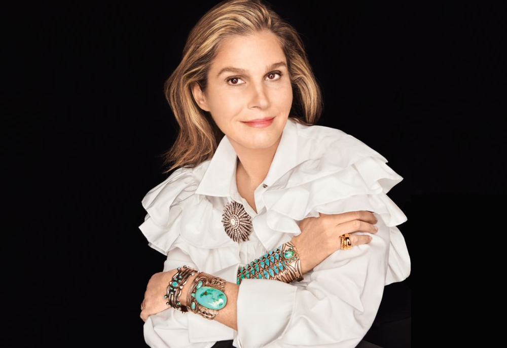 A woman with shoulder-length blonde hair wearing a white ruffled blouse and multiple bracelets, posing against a black background.