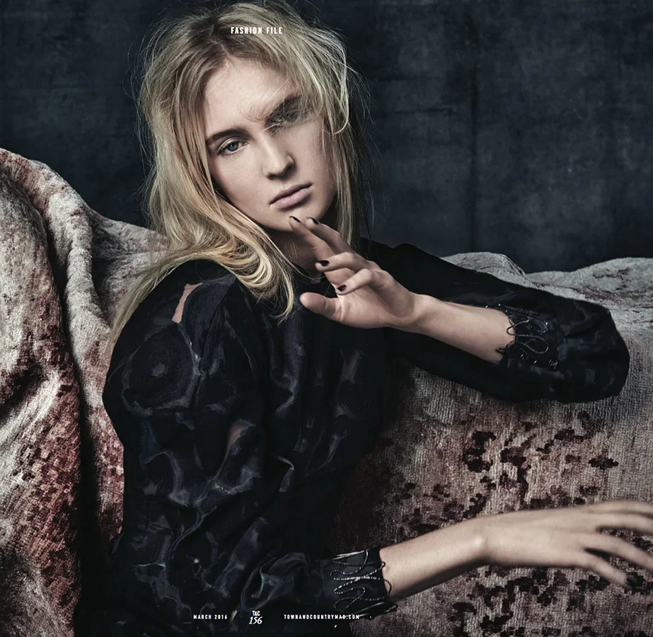 A young woman with blonde hair and a black top sitting on a textured sofa against a dark background, looking at the camera with her hand near her face.
