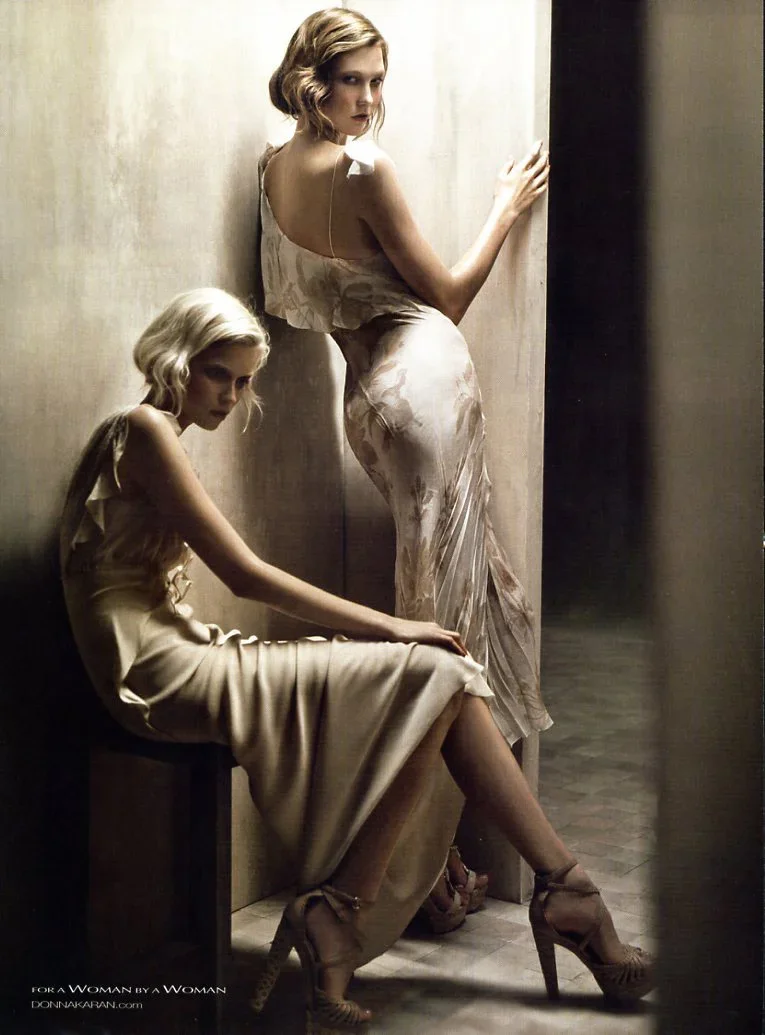 Two women with blonde hair in elegant dresses, one sitting on a bench and the other standing, in a dimly lit room with neutral tones.