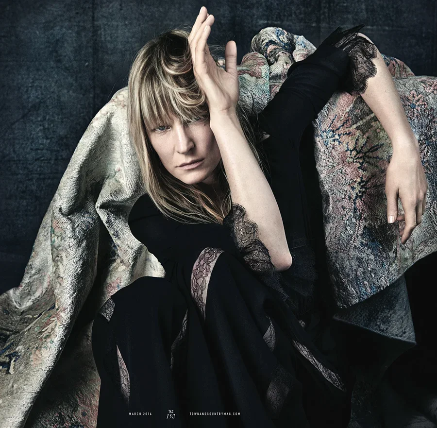 A woman with blonde hair and bangs, wearing a black dress with lace details, poses lying on a patterned and textured fabric surface, with one hand on her forehead and gazing at the camera.