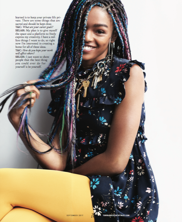 A young woman with multicolored braids smiling and posing for a photo. She is wearing a dark floral dress and has layered necklaces and jewelry. There is a white background with some text and a yellow object in the lower part of the image.