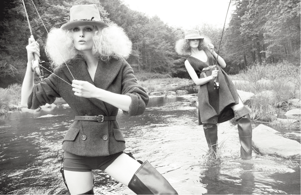 Two women with curly hair wearing big hats, boots, and stylish clothing, standing in a shallow river holding fishing rods in a natural outdoor setting with trees.