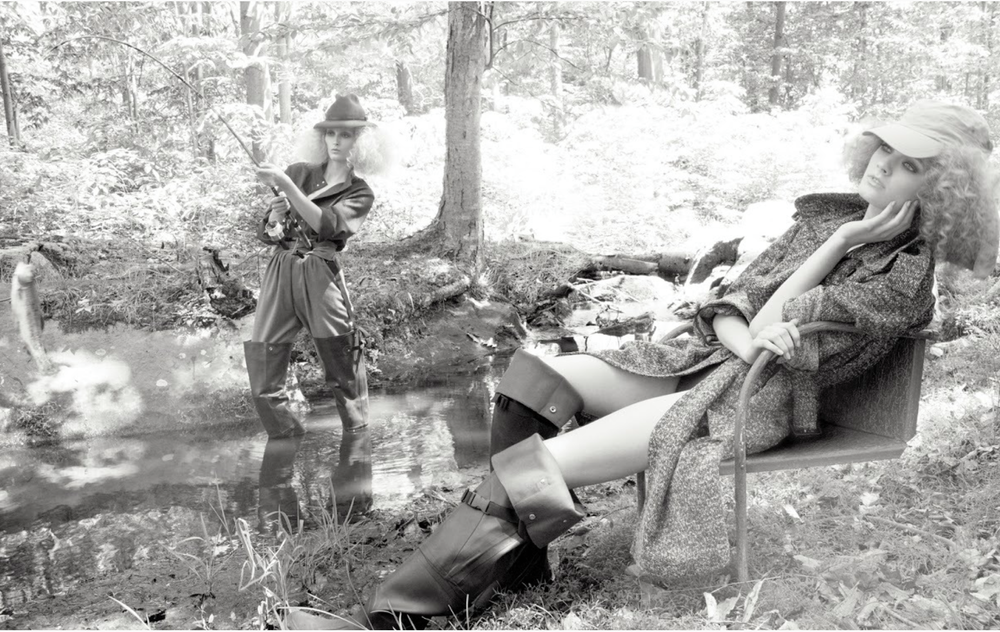 Two women in outdoors clothing, one sitting on a bench near a stream, and the other standing in the stream with a fishing rod in a forest setting.