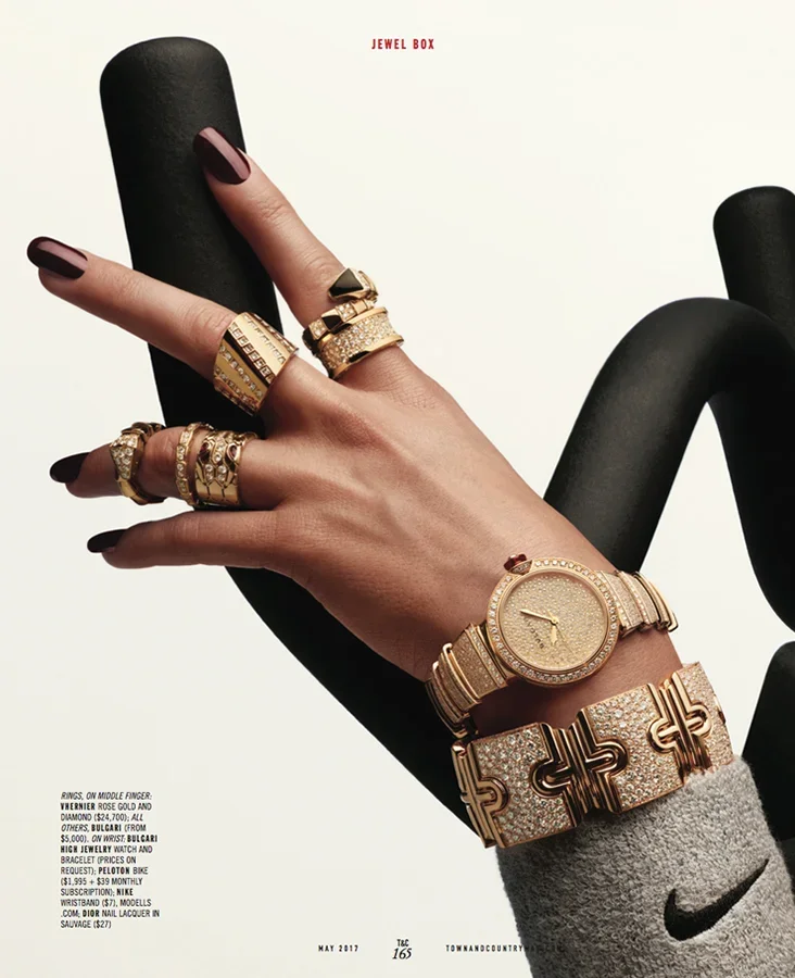 Close-up of a person's hand and wrist adorned with multiple gold and diamond jewelry pieces, including rings, a watch, and bracelets. The hand is holding a black object against a plain background.