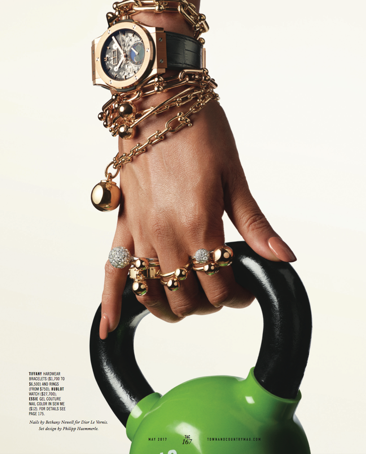 Close-up of a hand gripping a black and green kettlebell, adorned with multiple gold and jeweled rings and several gold bracelets, including a large watch with a leather strap, set against a plain white background.