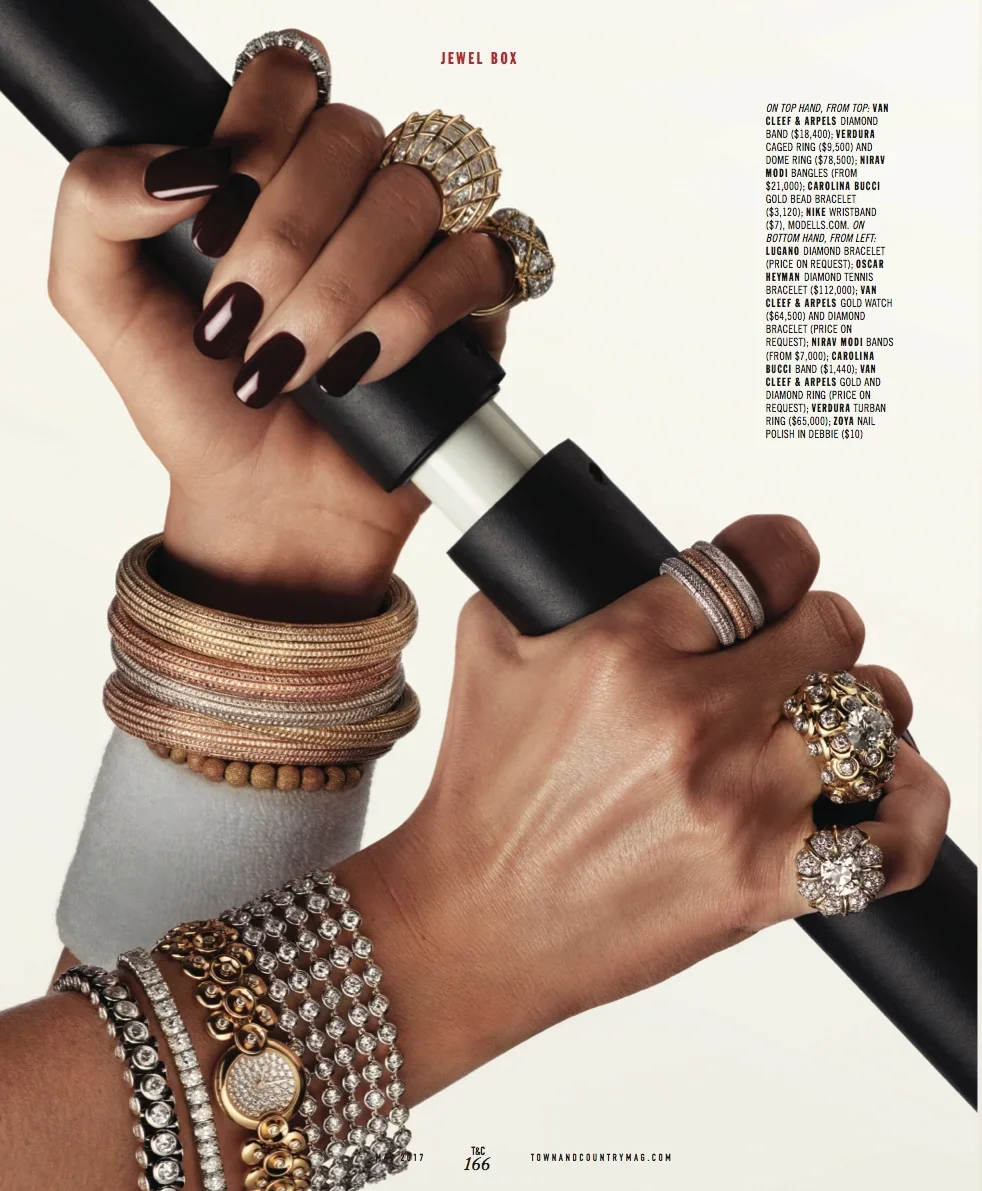 Close-up of hands adorned with various rings and bracelets holding a tube of lipstick, with a plain white background.