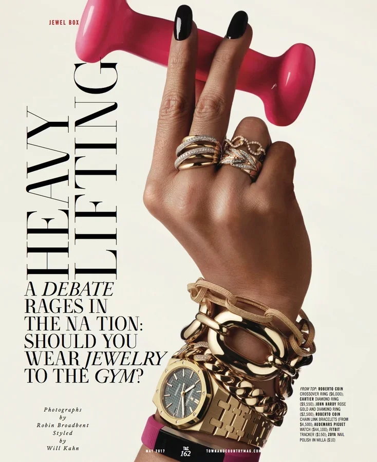 A hand with black nail polish holding a pink vintage rotary phone against a white background. The hand is adorned with multiple gold and silver rings and a gold watch with a gray face. Jewelry and accessories take up most of the focus, with text indi