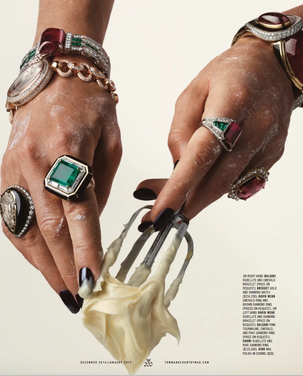 Close-up of hands adorned with various rings and bracelets, holding a cheese grater with cheese. The hands are dusty with flour or powdered sugar, and the jewelry includes emerald and ruby rings, emerald and diamond bracelets, a gold and diamond watc