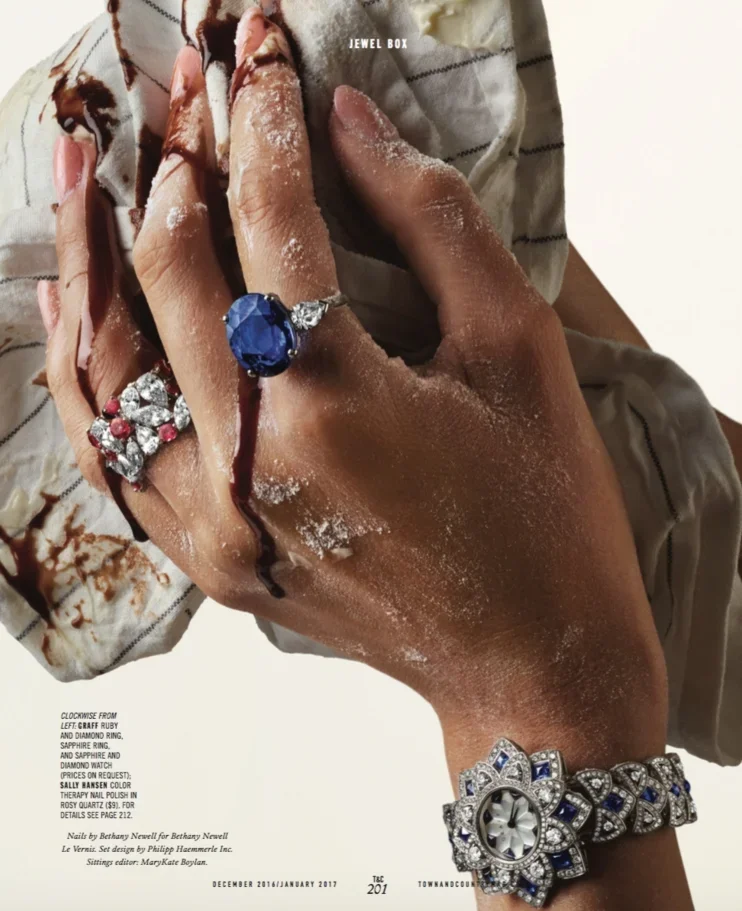 Close-up of a person's hands adorned with large jewelry rings and a bracelet, with some powder or chalk on their fingers and wrist, holding a piece of cloth with a floral pattern.