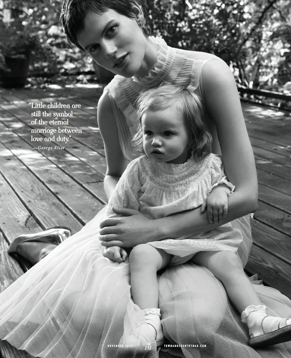 A woman with short hair sitting outdoors on a wooden deck, holding a young girl in her lap. The woman is smiling and looking at the camera, while the girl has a serious expression. The woman is wearing a sleeveless top with ruffles at the collar, and