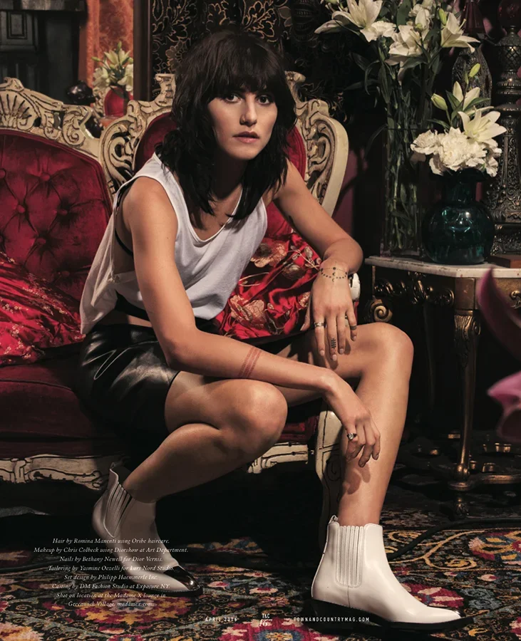 Young woman with black hair and bangs sitting on an ornate vintage-style sofa, wearing a sleeveless top, dark leather shorts, and white Chelsea boots, surrounded by an elegant vintage interior with floral arrangements.