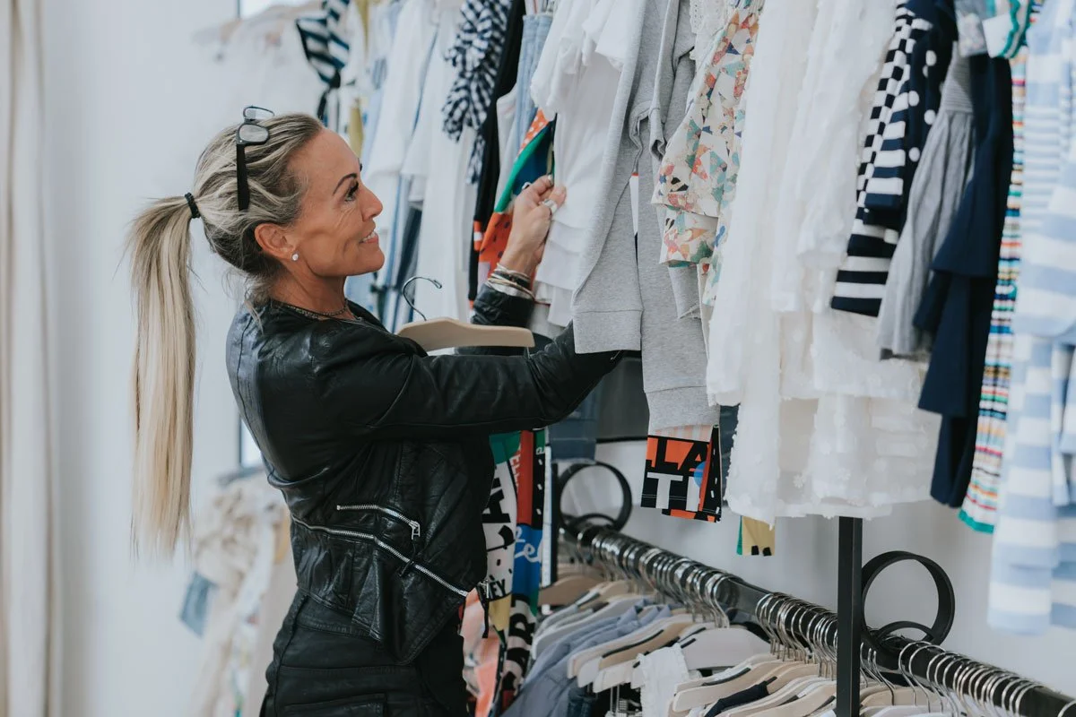 A woman shopping for clothes, smiling while browsing through a clothing rack filled with various shirts and blouses.