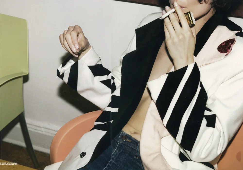 Person with curly hair wearing a patterned black and white blazer, a pink shirt with a red pocket, and jeans, holding a cigarette near their lips.