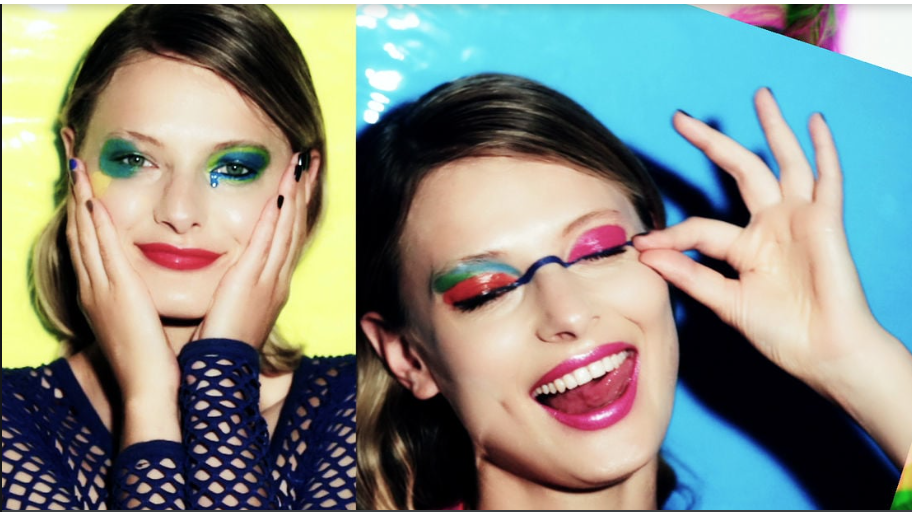 Two women with colorful makeup pose playfully against vibrant backgrounds. The woman on the left has green and blue eye makeup and red lipstick, while the woman on the right has pink and blue eye makeup, bright pink lipstick, and is holding glasses n