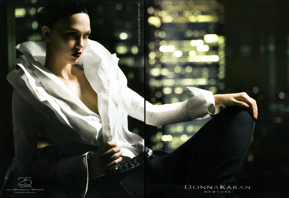 Fashionable woman wearing a white blouse with ruffles, sitting in a dimly lit room, nighttime cityscape visible through the window behind her.