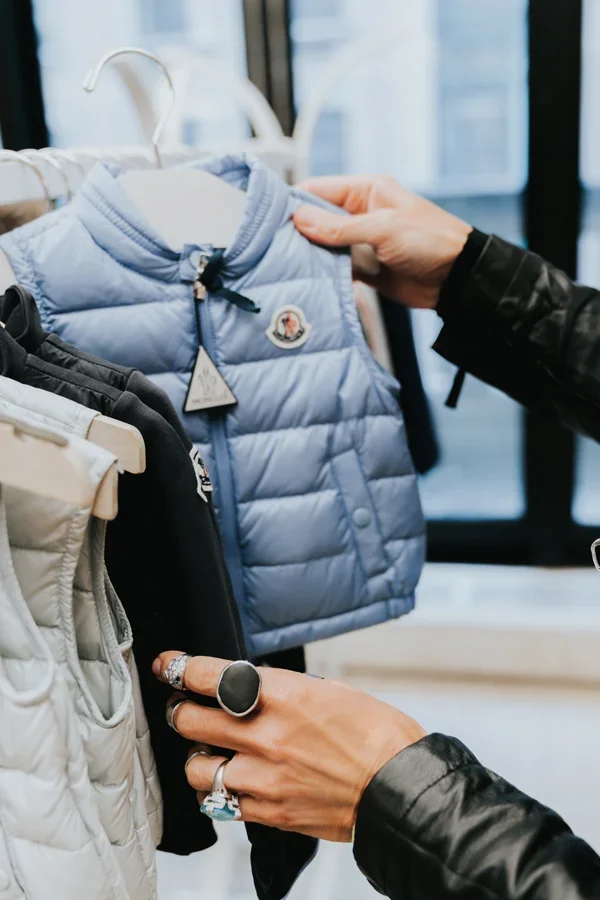 Person shopping for baby jackets in a clothing store, touching a light blue padded jacket with a Moncler logo.