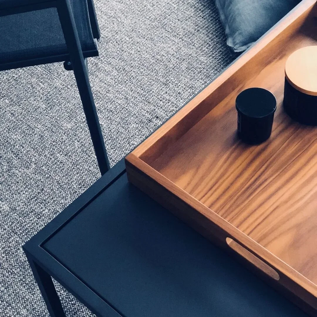 Close-up of a wooden tray on a black table, with two black and beige containers inside. Part of a grey chair and a textured carpet are visible in the background.