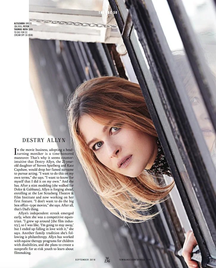 A woman with blond hair and blue eyes leaning against a black metal fence, with a magazine article visible in the background.