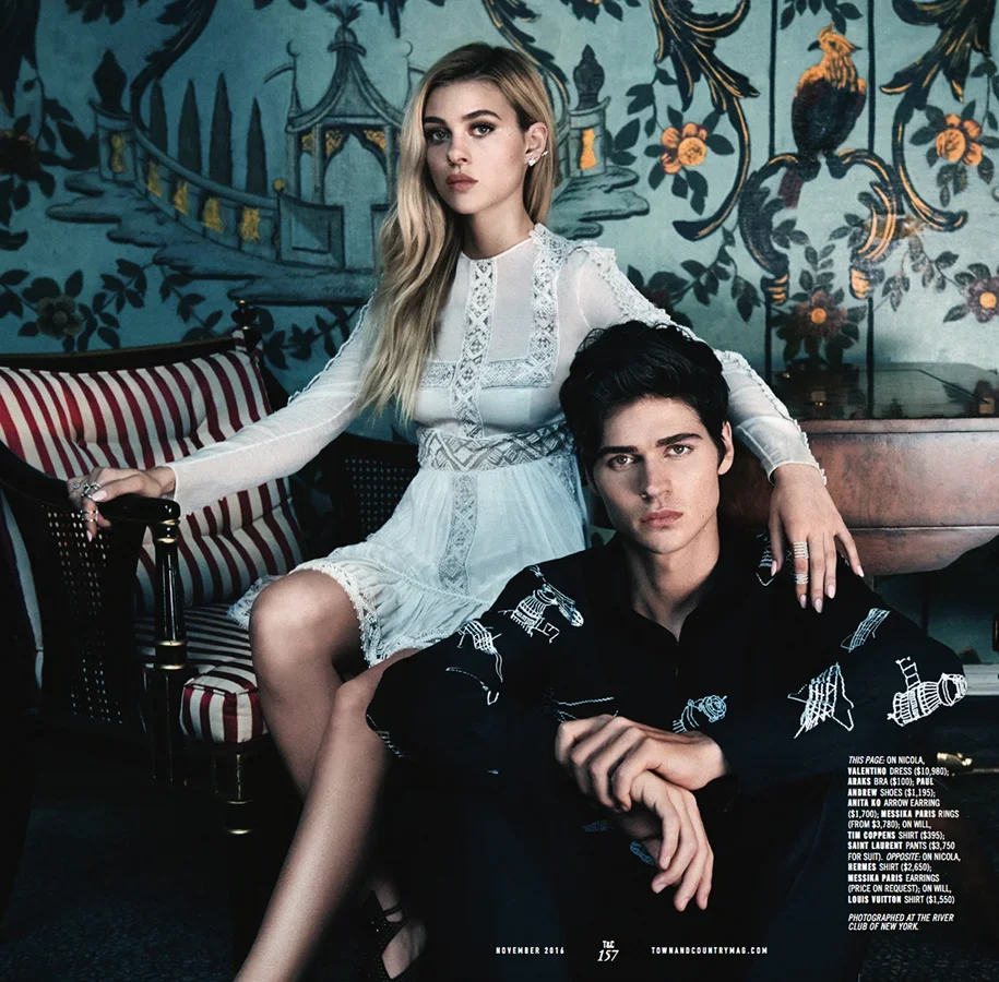 A young woman with long blonde hair wearing a white lace dress sits on a vintage-style striped sofa, with her arm resting on a young man with black hair who is dressed in a black shirt with white animal motifs, seated in front of her, on a richly dec