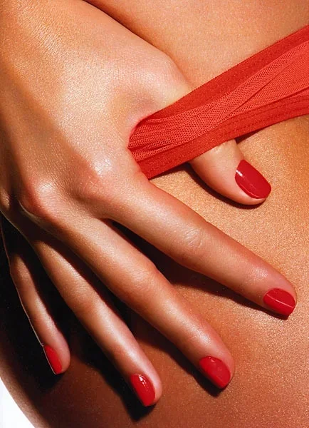 Close-up of a woman's hand with red fingernail polish gently touching her skin, with red fabric in the background.
