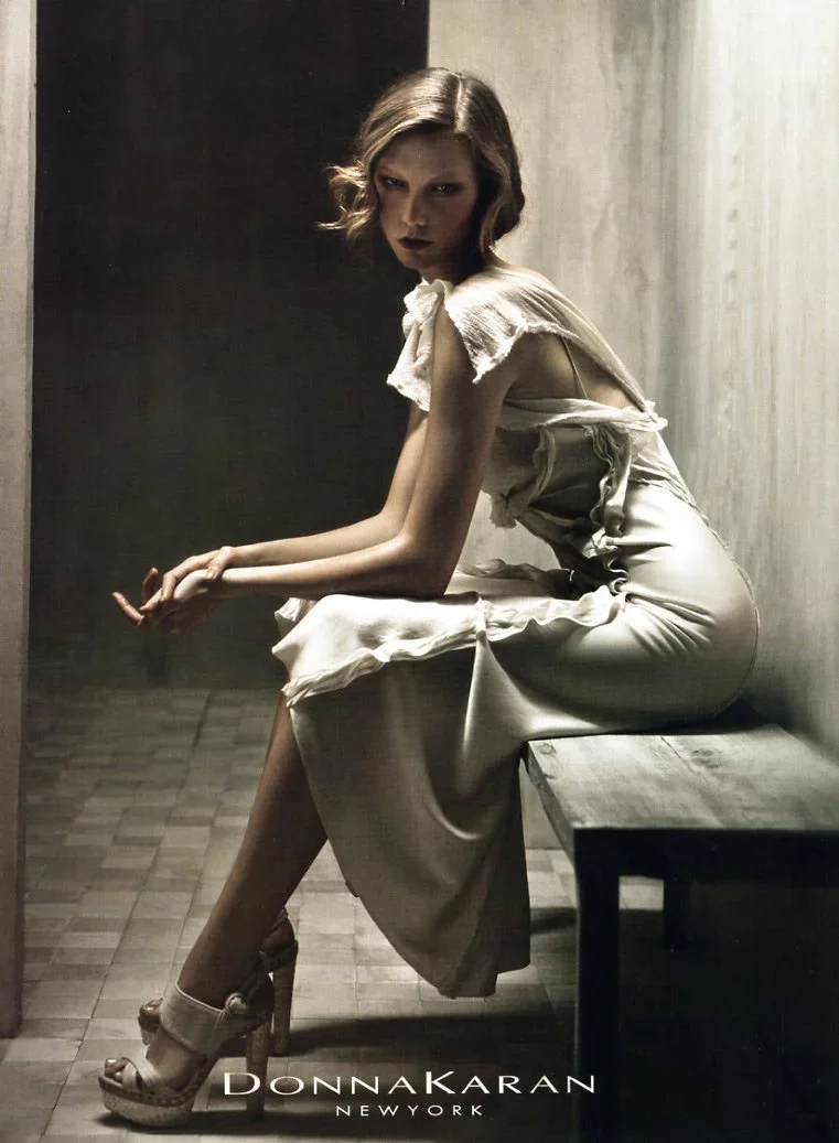 A woman with wavy hair sitting on a wooden bench, wearing a light-colored dress and high heels in a dimly lit room.