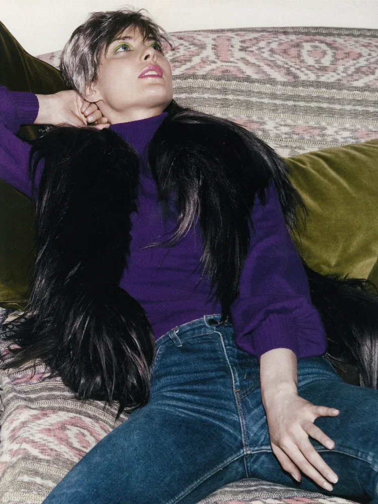 A woman with long dark hair and light skin is lying on a couch, wearing a purple top and jeans. She has one hand resting on her thigh and the other behind her head, looking upwards. The couch has patterned and plain pillows.
