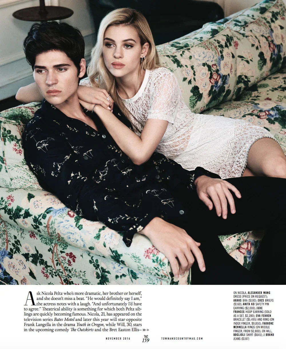 A young man and woman lying on a floral-patterned sofa, with the woman leaning on the man's shoulder. The young man has dark hair and is wearing a black shirt with a light pattern. The woman has blonde hair and is wearing a white lace dress.