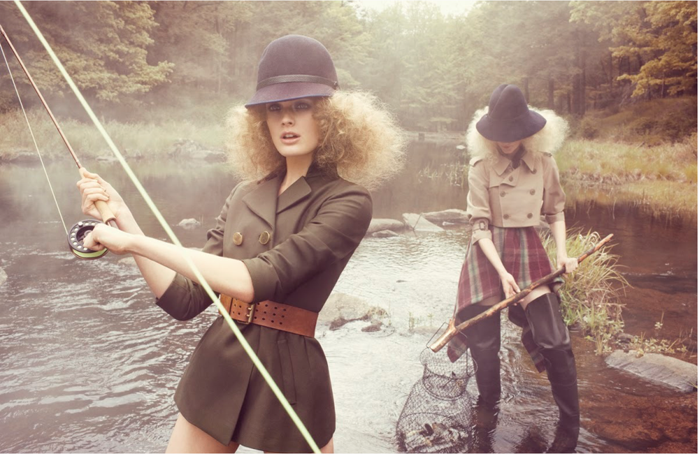 Two women dressed in vintage-style clothing fishing in a river surrounded by trees.