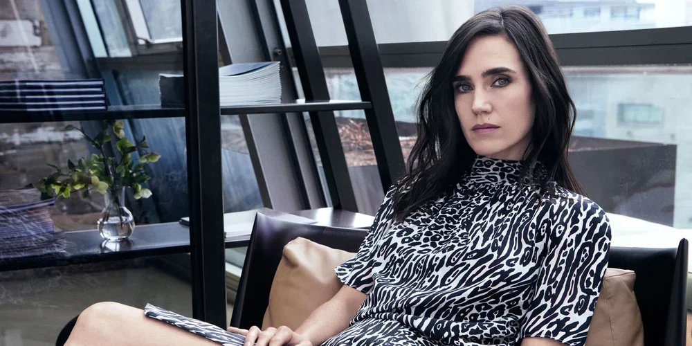 A woman with dark hair and makeup, wearing a black and white animal print dress, sitting on a beige sofa in a modern, well-lit indoor space with large windows and shelves.
