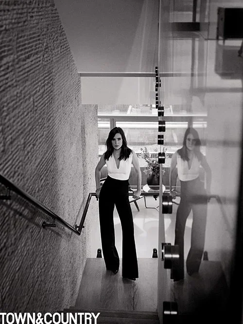 A woman with dark hair standing on a staircase near a large window, with her reflection visible on a glass wall to her right, in a modern indoor setting.