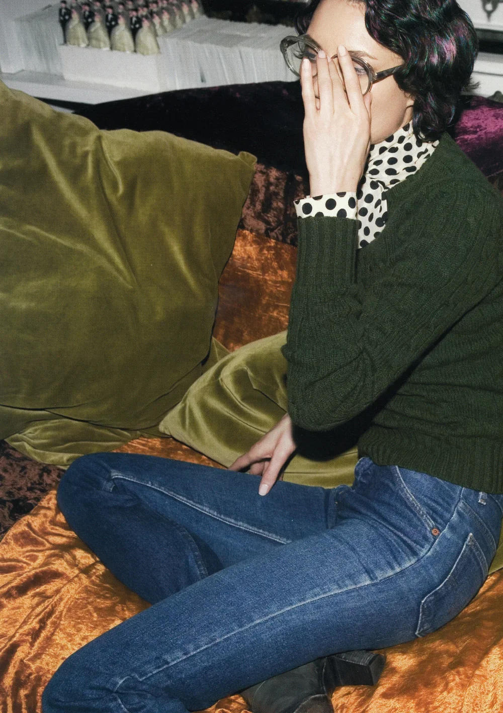 A woman with glasses and dark hair with colorful streaks sitting on an orange velvet couch, covering her face with her hand, wearing a polka dot shirt under a green sweater, and blue jeans.
