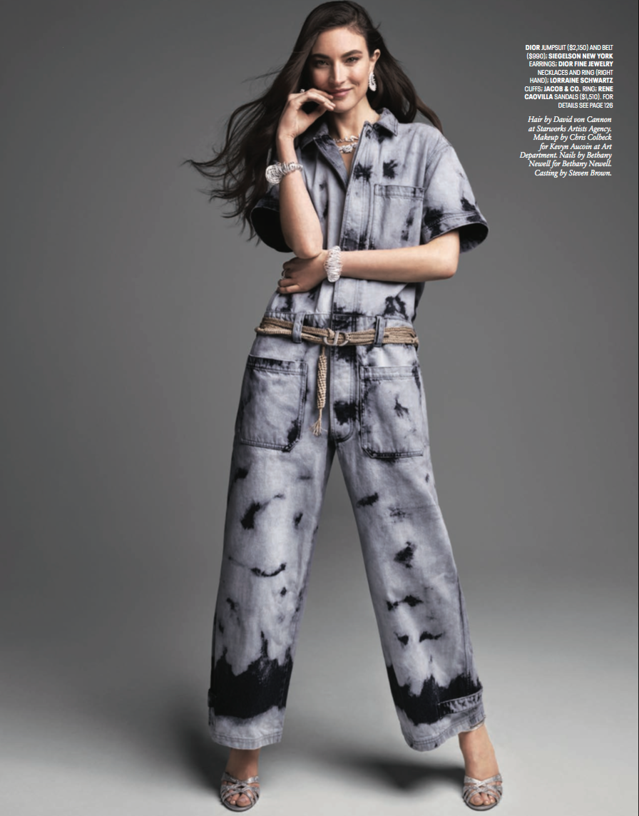 A woman wearing a silver and black diamond-printed denim jumpsuit, accessorized with silver jewelry including earrings, a necklace, a ring, a bracelet, and a watch, with silver high heels.