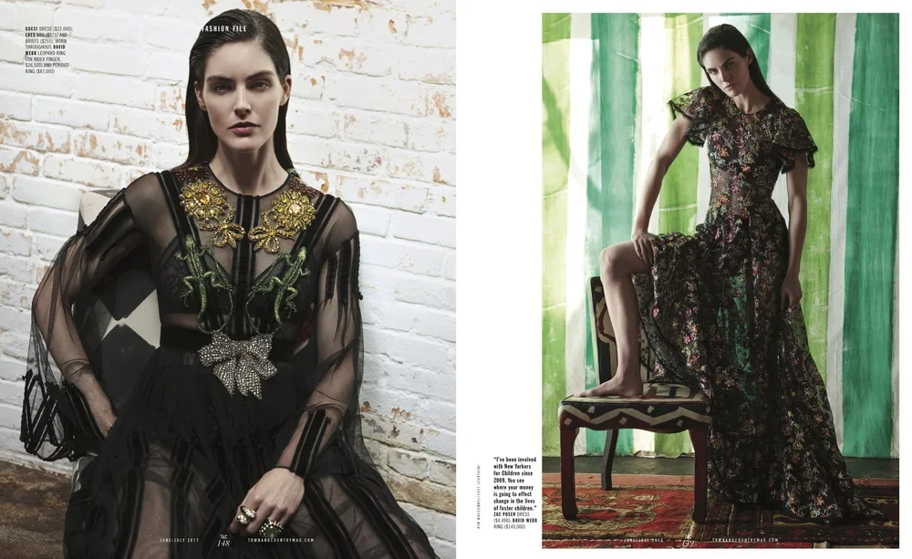 Two women modeling fashion outfits. The woman on the left is wearing a black sheer dress with floral embellishments and accessories, standing against a white brick wall. The woman on the right is sitting on a chair, wearing a long floral dress, with 