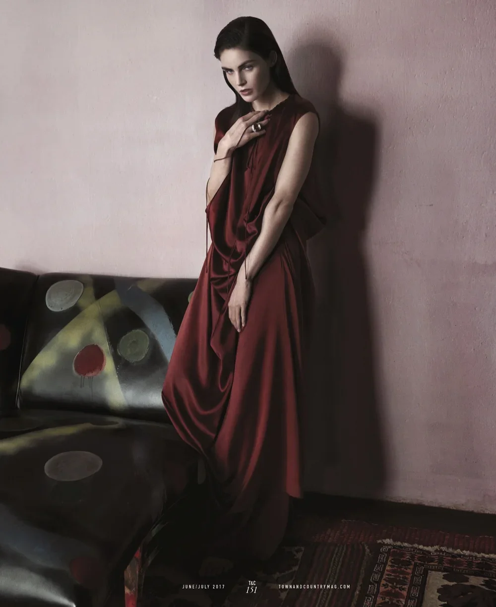 A woman in a flowing red dress standing in a room with pink walls, next to a black couch with colorful circular patterns.