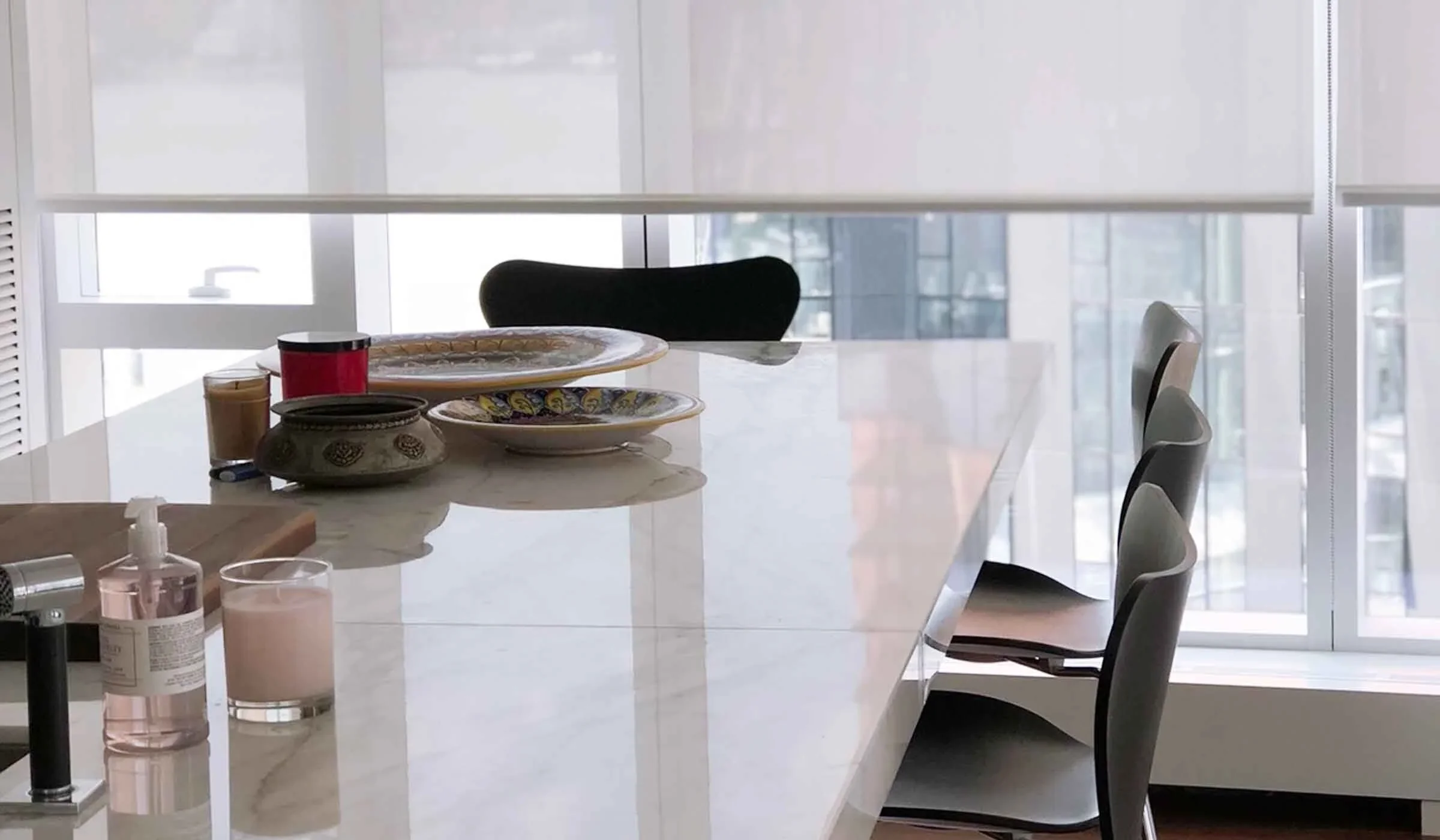 Empty dining table with decorative dishes, candles, hand sanitizer, and beverages in a modern room with large windows and city views.