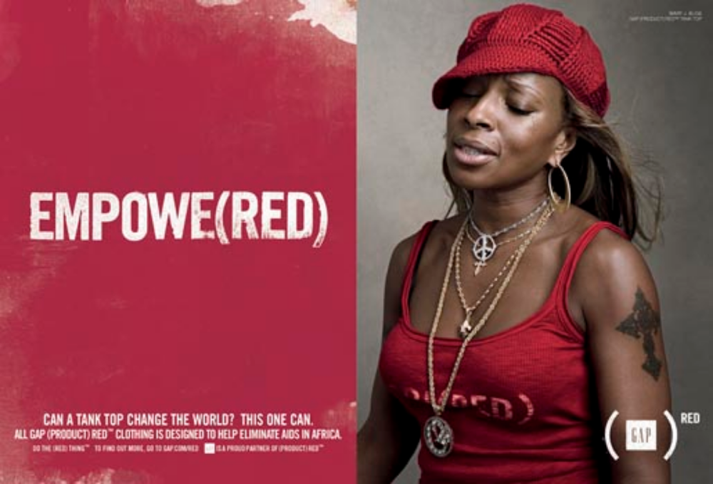 A woman wearing a red tank top and a red hat, with gold jewelry, including earrings and layered necklaces, and a tattoo on her arm. Next to her is a pink poster with the text "EMPOWER(RED)" and a quote about changing the world.