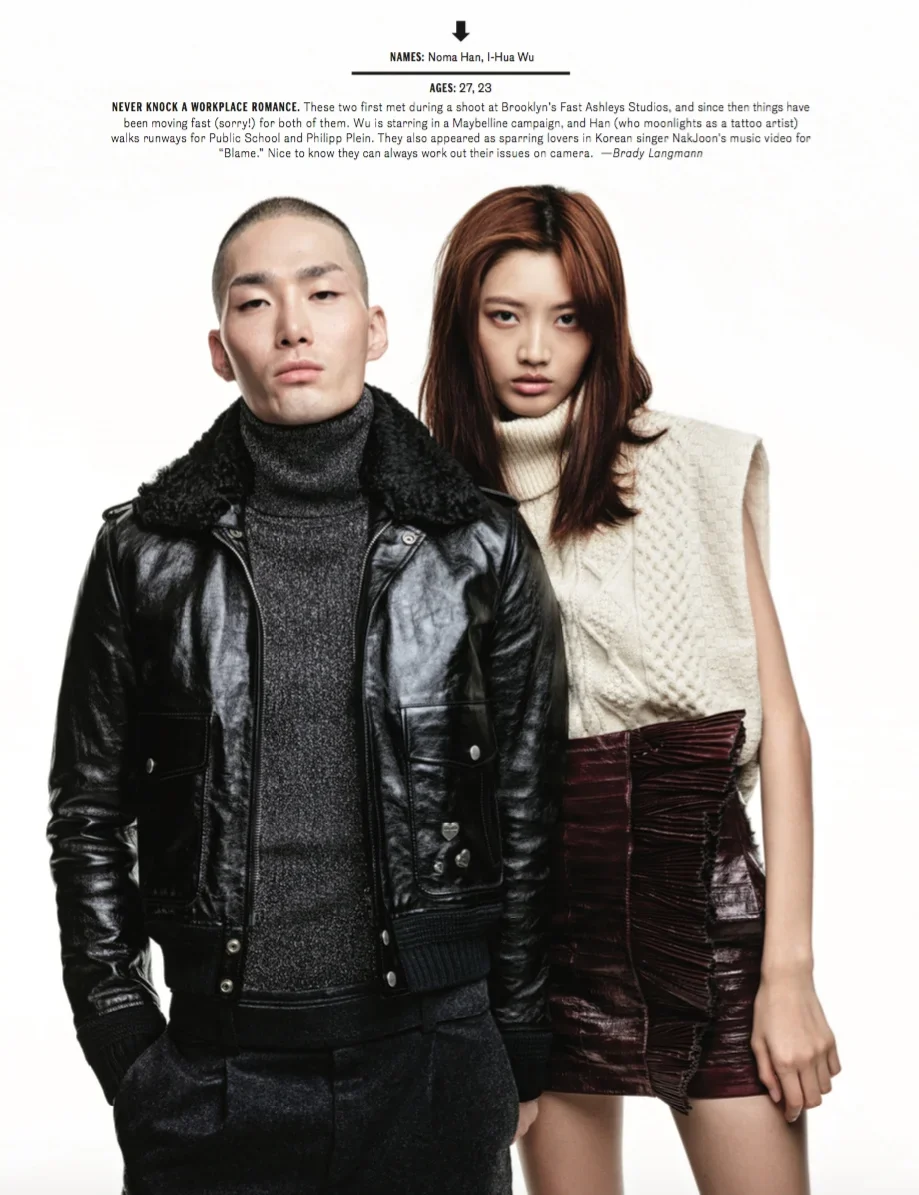 A young man and woman stand side by side against a plain background. The man has a shaved head and wears a black leather jacket over a gray sweater, with hands in his pockets. The woman has long, wavy brown hair and wears a sleeveless beige knit top 