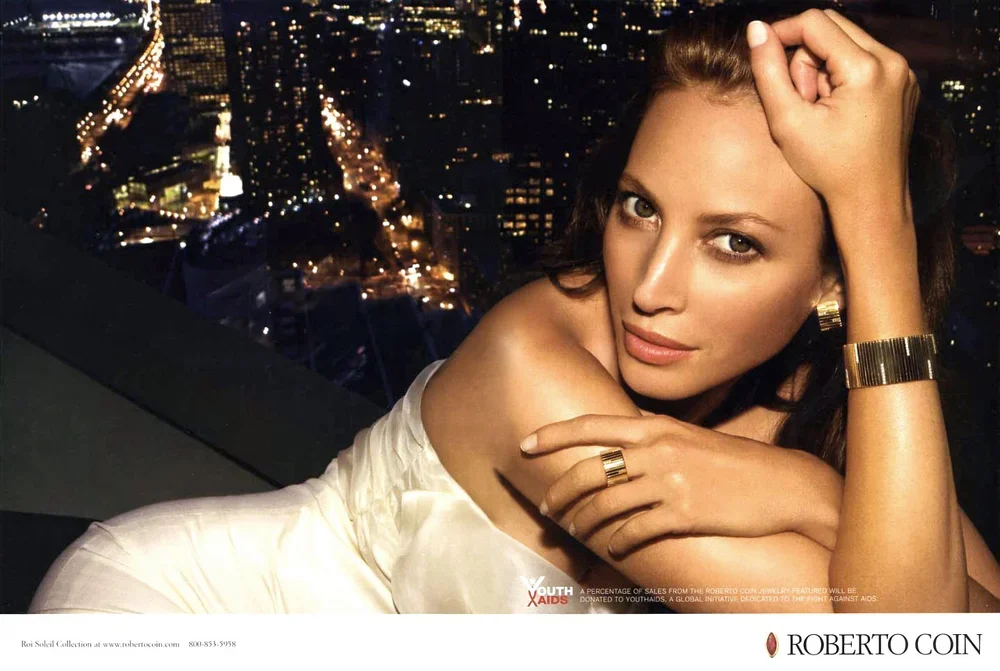 A woman with brown hair and light skin, wearing gold jewelry including a bracelet, ring, and watch, is leaning on her arm and looking into the camera with a cityscape illuminated with lights in the background.