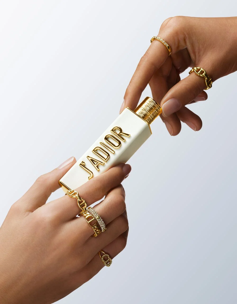Two hands holding a white and gold perfume bottle labeled 'J'ADIOR' with gold jewelry on both hands.