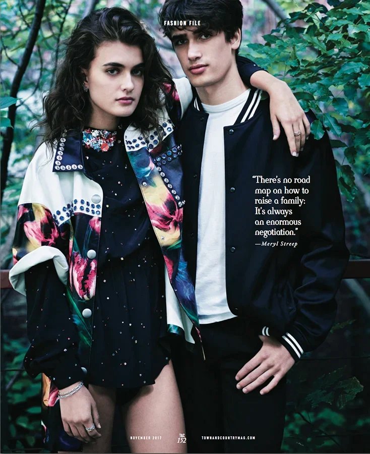 A young woman and man pose together outdoors, with trees and leaves in the background. The woman wears a colorful floral jacket over a dark dress, and the man wears a black and white jacket over a white shirt. The woman has curly dark hair, and the m