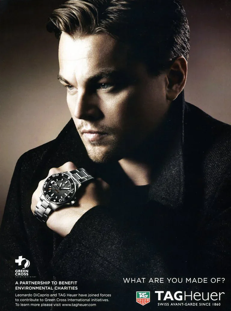 Portrait of a man with slicked-back hair wearing a black jacket, showing a silver TAG Heuer watch on his left wrist, with a serious expression and looking to the side.