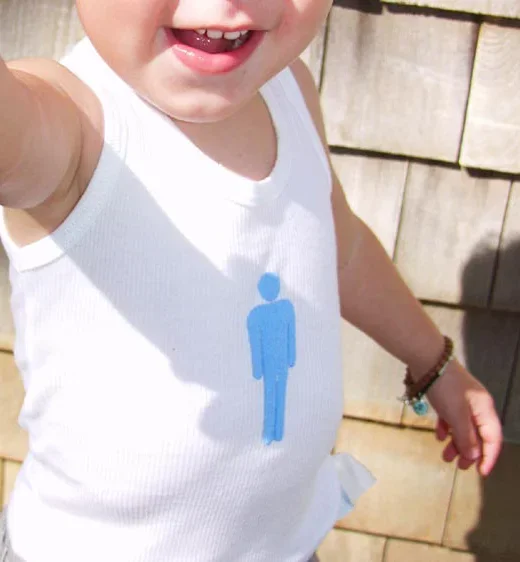 Close-up of a smiling child wearing a white tank top with a blue gender symbol and a beaded bracelet, outdoors with wooden fencing in the background.