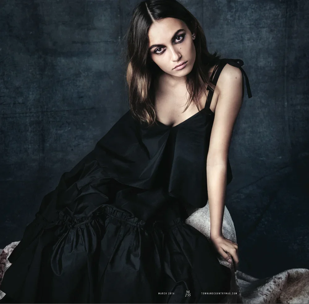 A woman with dark hair and smoky eye makeup, wearing a black dress, sitting against a dark textured backdrop.