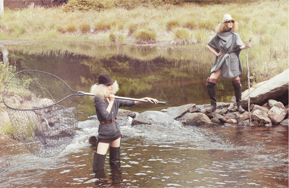 Two women in outdoor clothing standing in a river, one holding a fishing net and the other holding a walking stick, with grassy landscape in the background.
