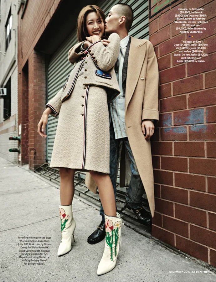 Two fashionable young adults, a woman and a man, leaning against a brick wall in an urban setting. The woman is smiling, wearing a beige coat and white embroidered cowboy boots, while the man is dressed in a beige overcoat with layered casual clothin