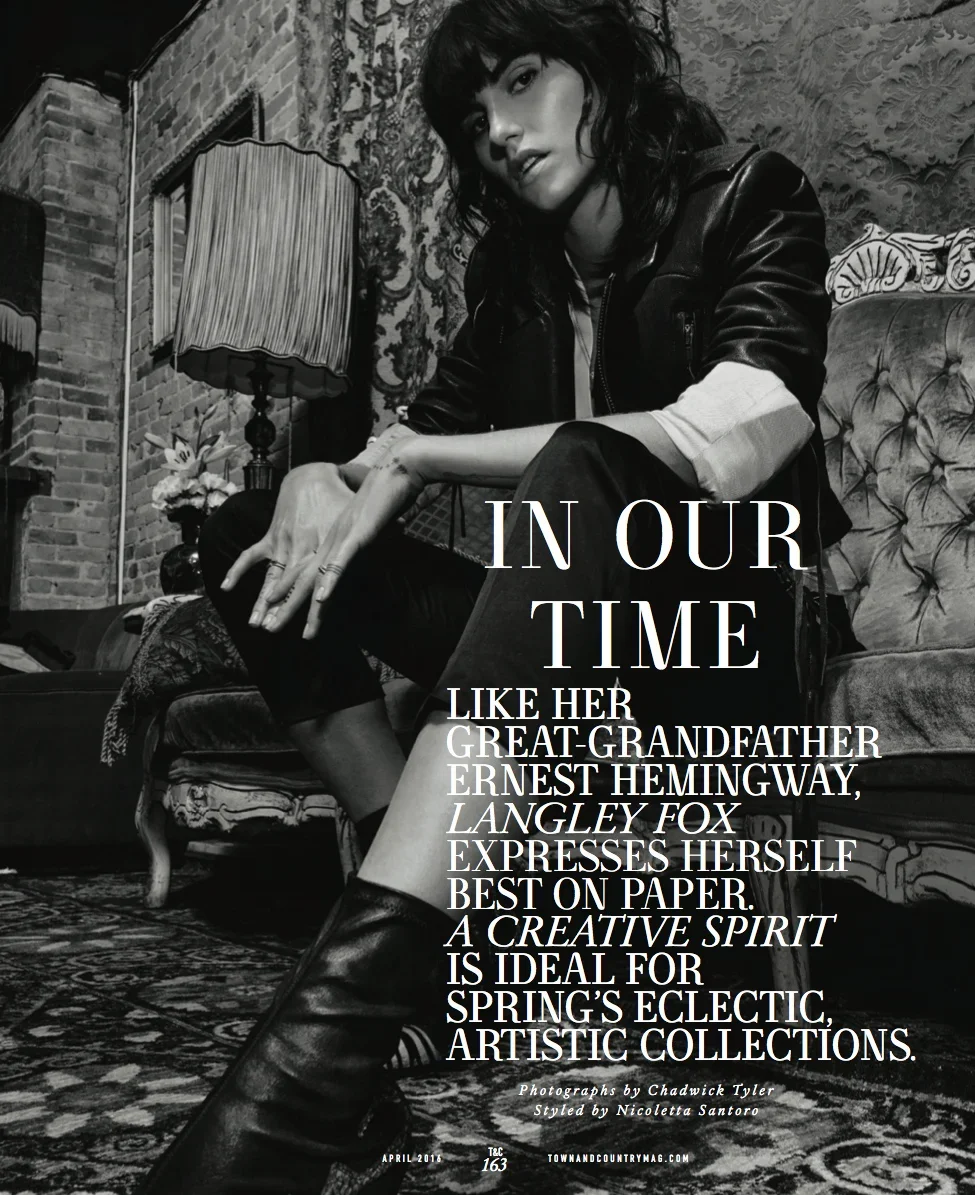 Black and white photograph of a woman with dark hair sitting on a vintage-style sofa in a cozy, artistically decorated room. She is wearing a leather jacket, with a confident pose and expression. There is a lamp, a flower arrangement, and ornate wall